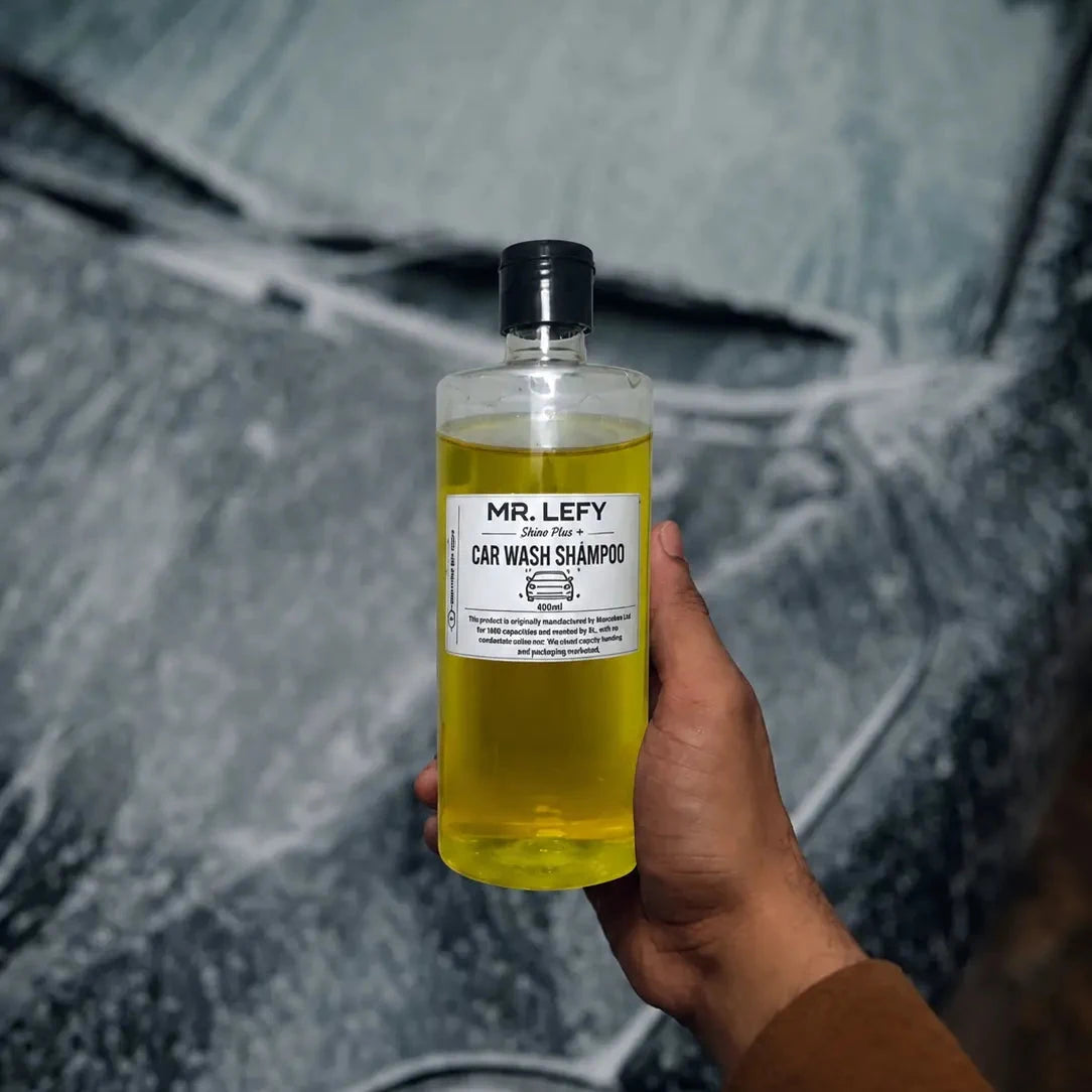 A hand holding a clear bottle of Mr. Lefy Car Wash Shampoo filled with yellow liquid, set against a blurred gray car background.