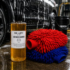 A bottle of Mr. Lefy Silver Mist Car Wash Shampoo stands next to two microfiber cleaning mitts, one red and one blue, in front of a wet car with soap foam in a garage setting.
